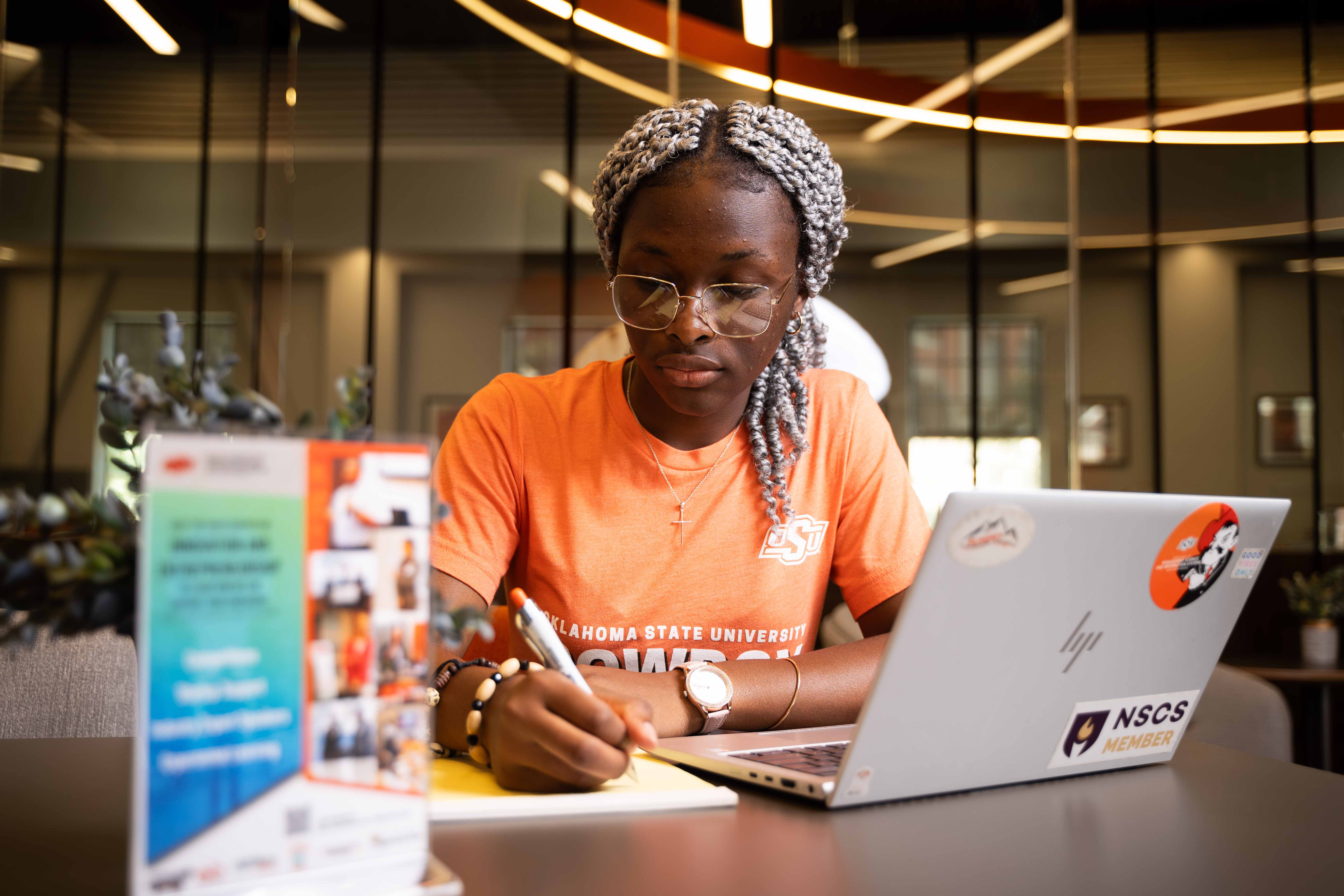 Female student wearing an OSU t-shirt writing notes with a laptop next to her.