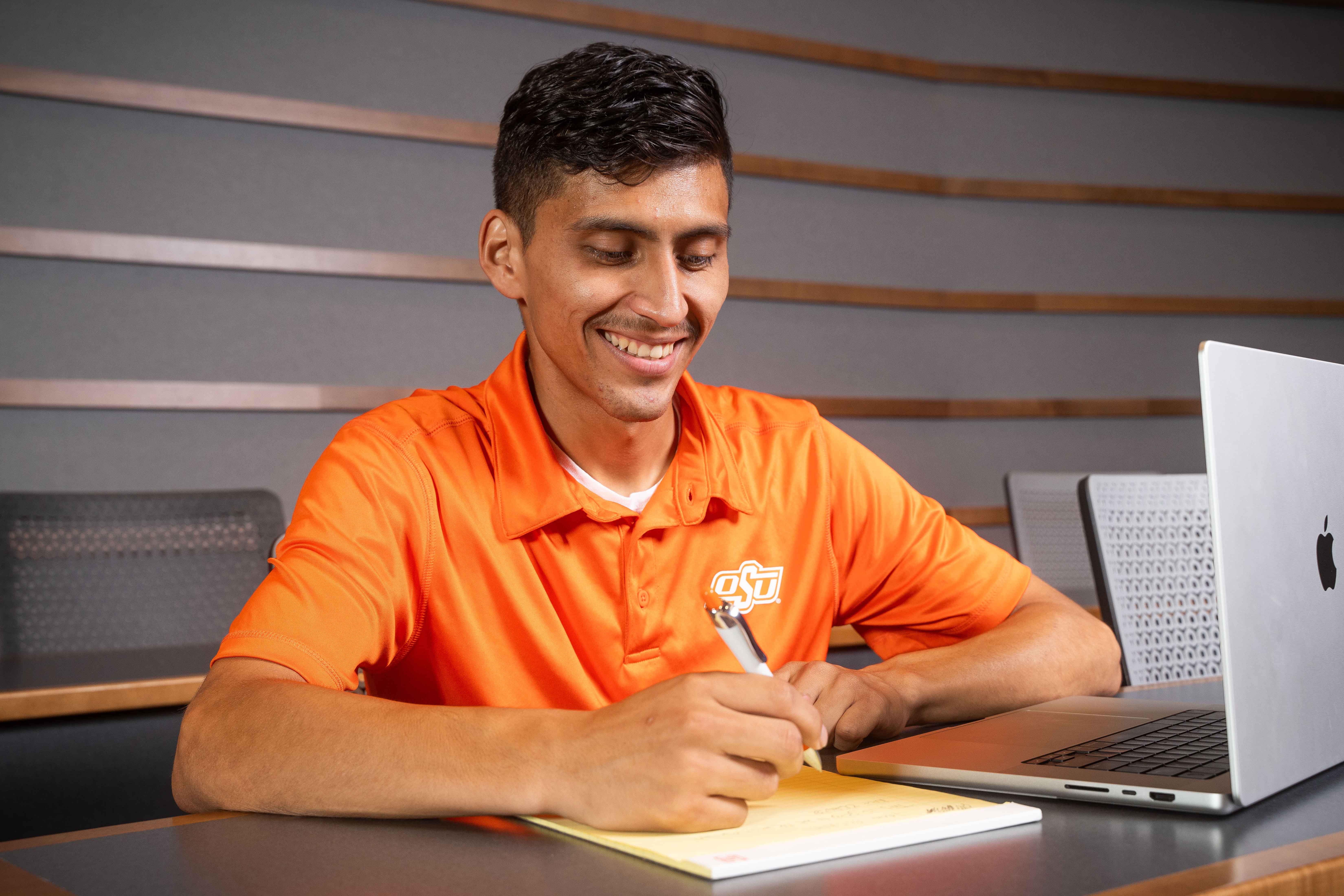 Male student wearing an orage OSU polo shirt taking notes with a laptop beside him. Male student wearing an orage OSU polo shirt taking notes with a laptop beside him.